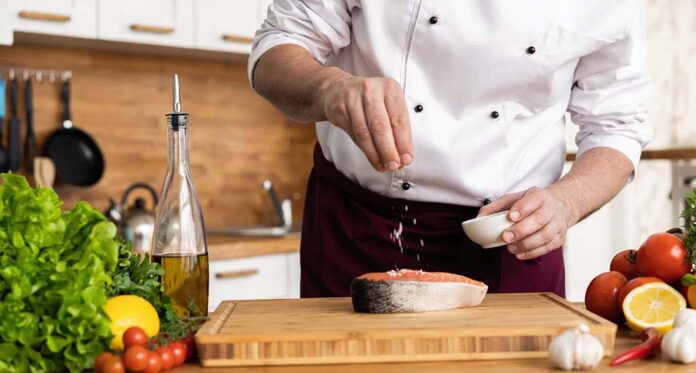 The chef prepares fresh fish salmon, trout, sprinkles with sea salt and vegetables. Horizontal photo. Concept cooking healthy and vegan cuisine, clean food, restaurants, hotel business.
